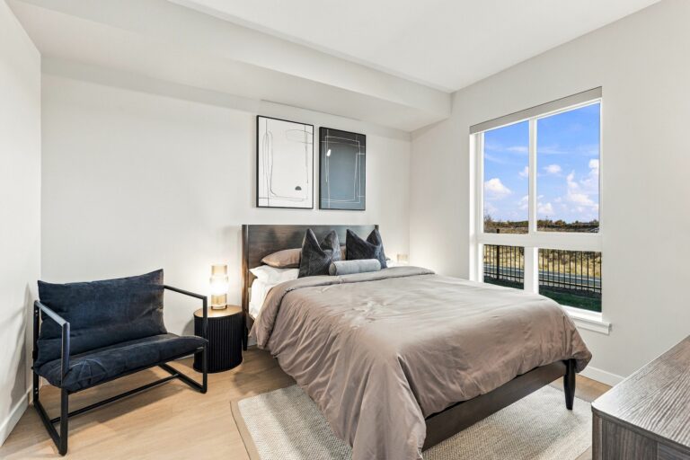 Furnished bedroom in a Bob Apartments unit, with a large window, queen bed, lounge chair, and contemporary decor