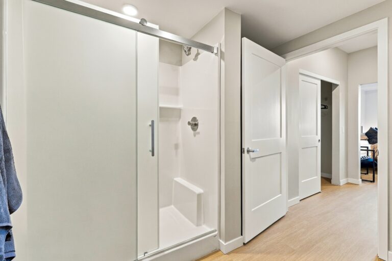Bathroom in a Bob Apartments unit with a sliding glass shower door, white tile surround, and hallway leading to a bedroom